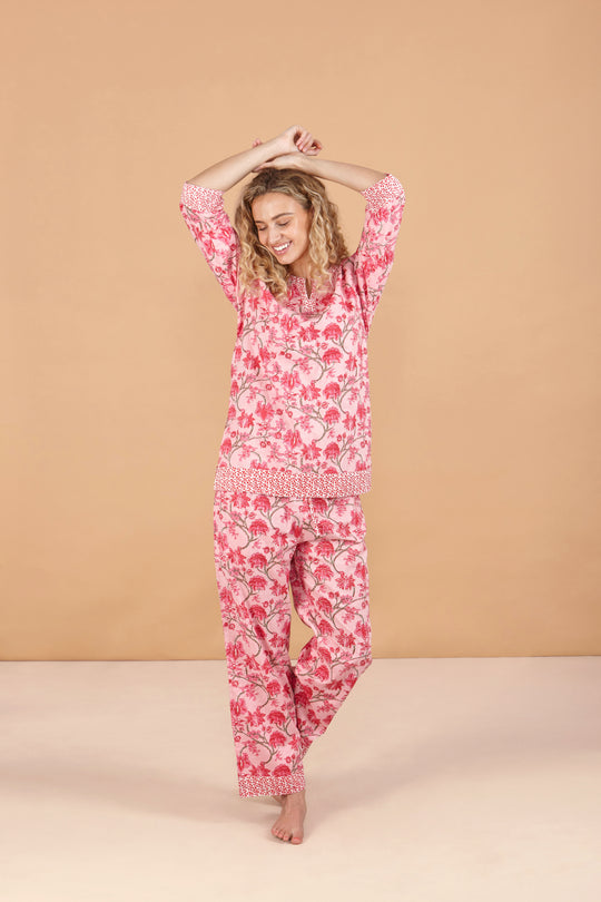 A woman standing with her hands raised, wearing a pink cherry blossom print pajama set with a split neckline and 3/4 sleeves, and matching pyjama bottoms with pockets.